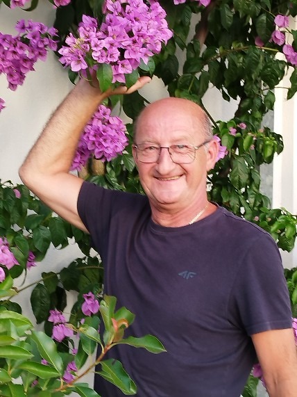 Uśmiechnięty mężczyzna w okularach pozuje przy kwitnącej bugenwilli, trzymając gałązkę z kwiatami w dłoni.