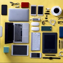 kompiki.pl Wojciech Rolak - Żółte tło z laptopami, smartfonami, słuchawkami, okularami, tabletem graficznym, klawiaturą, myszkami i innymi akcesoriami, ułożonymi w uporządkowany sposób.