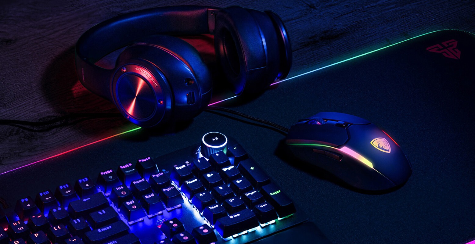Podświetlana klawiatura, mysz i słuchawki gamingowe na podkładce. Klawiatura mechaniczna z regulacją głośności. Sprzęt oświetlony w kolorach tęczy, na ciemnym tle.