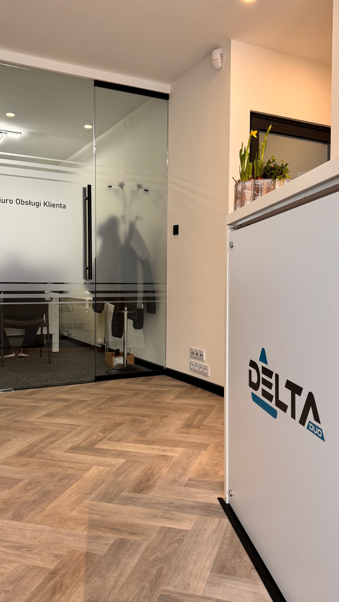 Nowoczesne biuro z recepcją Delta Duo, parkiet w jodełkę, przeszklone drzwi z napisem 'Biuro Obsługi Klienta' i rośliną w tle. Minimalistyczny design.