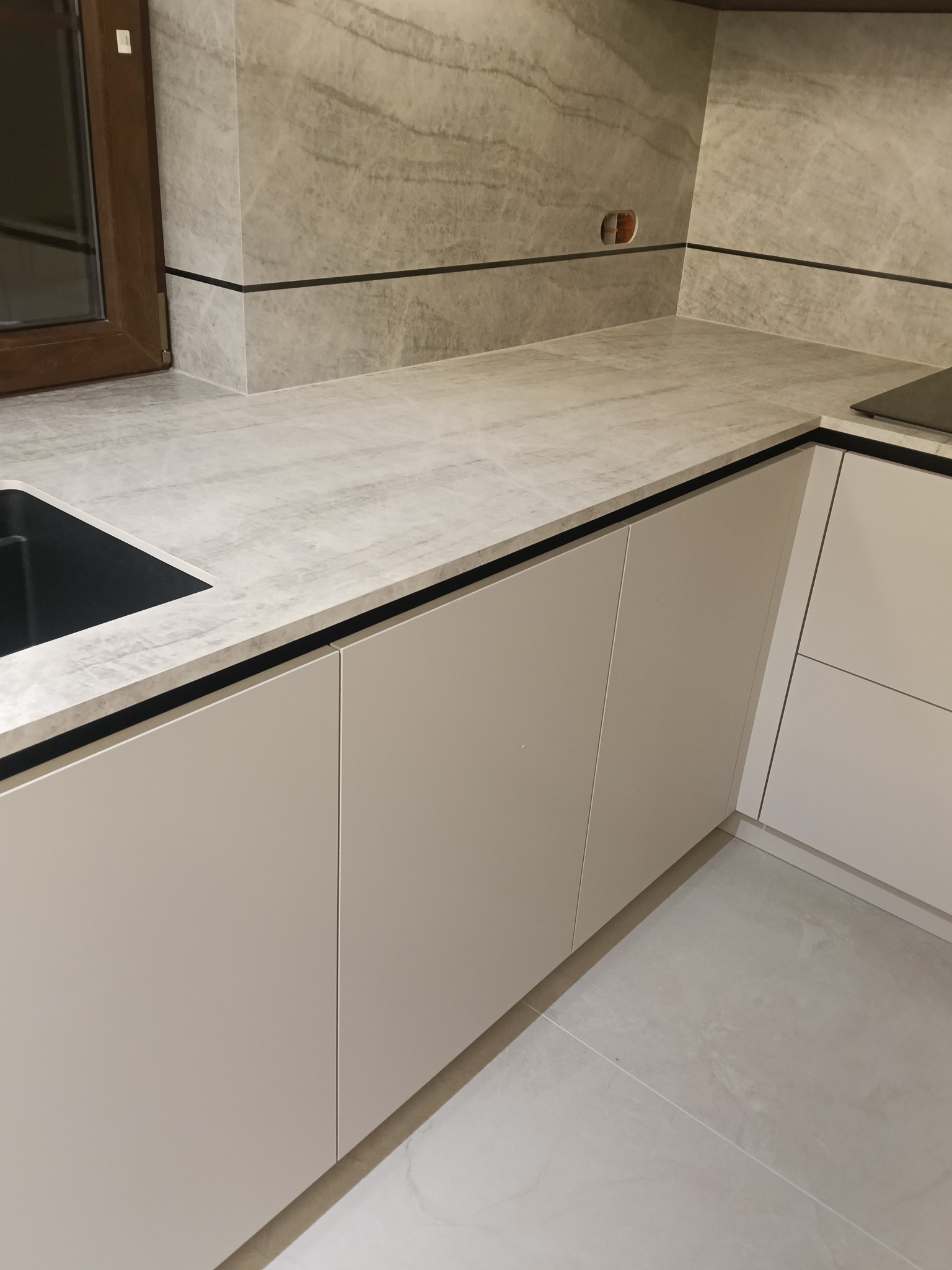 Jasna kuchnia z marmurowym blatem i płytkami. Widoczny fragment okna, zlew i płyta grzewcza. Minimalistyczny design z subtelnymi akcentami kolorystycznymi.