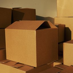 BGU Usługi Transportowe Przeprowadzki Transport Mebli Białystok - Stos kartonowych pudeł do pakowania, oświetlonych ciepłym światłem, z jednym otwartym pudłem na pierwszym planie, ukazującym gotowość do załadunku.