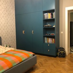 Jakub książek - Niebieska szafa wnękowa z półkami na książki i zabawkami w pokoju dziecięcym, widoczne skórzane uchwyty, parkiet w jodełkę i plecak szkolny obok.