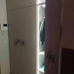 Jakub książek - Beżowa szafa z oświetleniem LED wewnątrz, ozdobne uchwyty w kształcie liści miłorzębu, widoczny fragment wnętrza z ubraniami i akcesoriami.