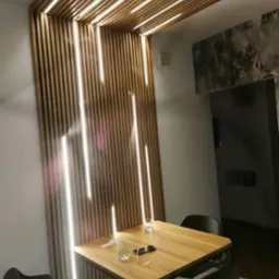 Drewniana ściana działowa z wbudowanym oświetleniem LED, tworząca dekoracyjny element w jadalni z kwadratowym stołem i czarnymi krzesłami.