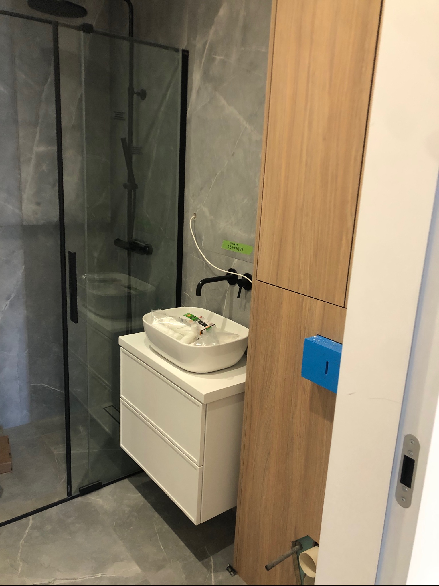 Nowoczesna łazienka w trakcie wykańczania: kabina prysznicowa z czarnymi ramami, biała umywalka nablatowa na szafce z szufladami, drewniana szafka wisząca z niebieskim podajnikiem, szare płytki...