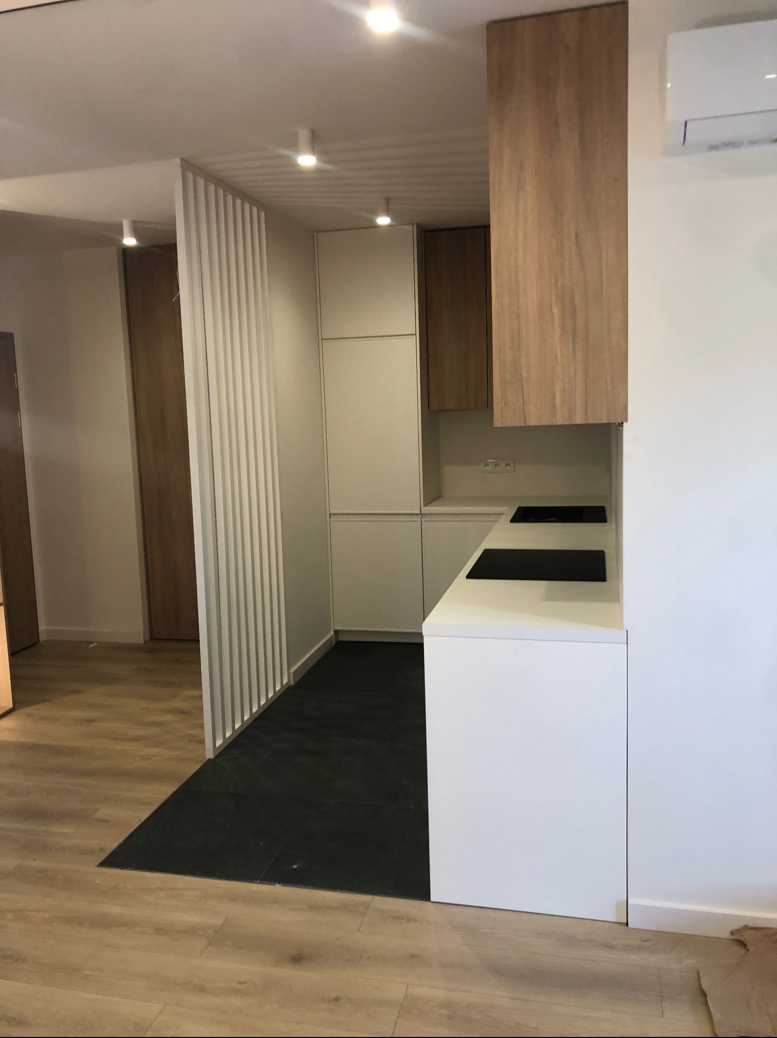 Nowoczesna kuchnia z białymi frontami i drewnianymi akcentami, widok na płytę indukcyjną w białym blacie, z podziałem przestrzeni za pomocą białych pionowych listew.