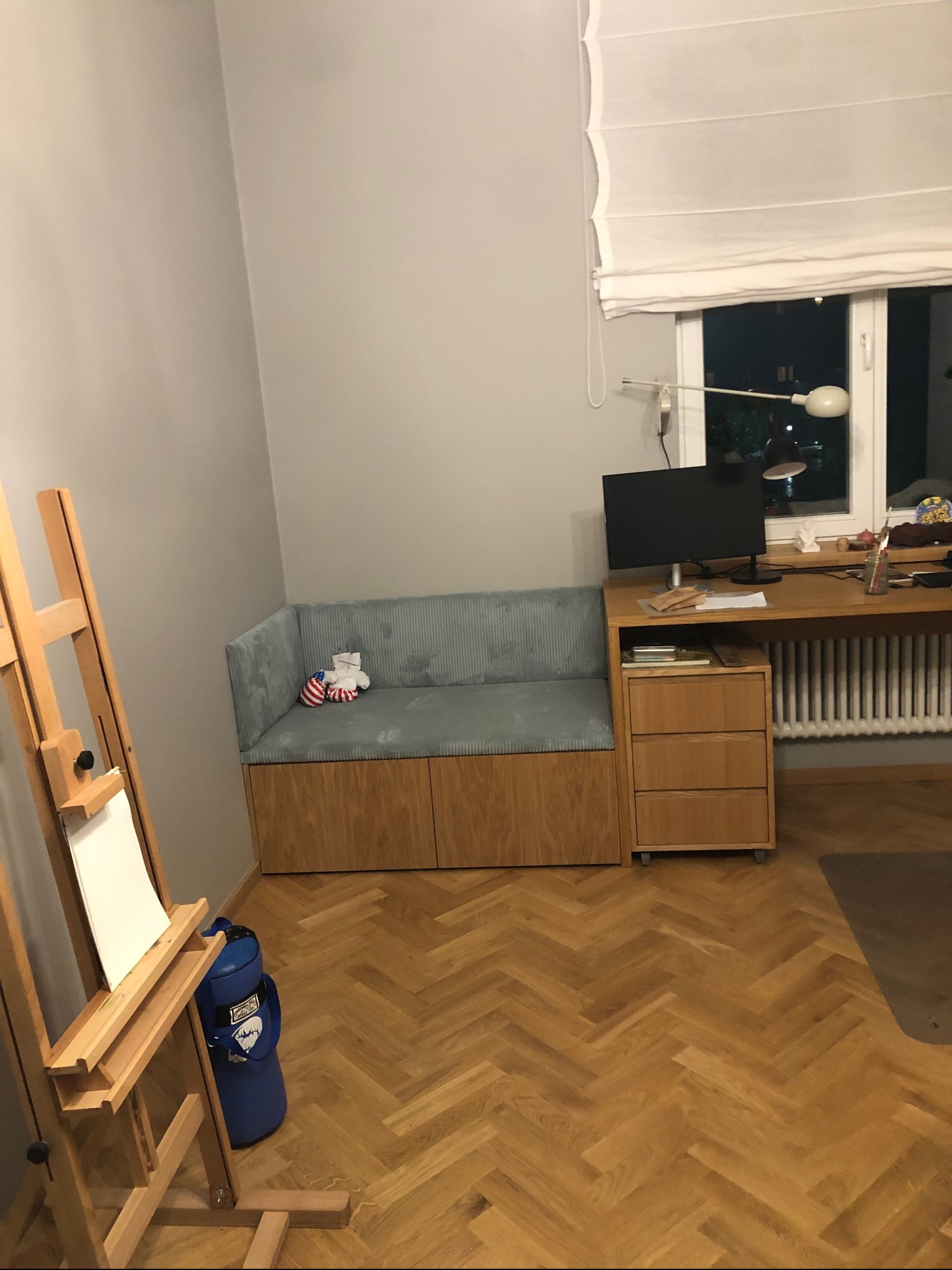 Kącik w pokoju z drewnianą zabudową siedziska z tapicerowanym, szarym siedziskiem, obok drewniane biurko z monitorem, na podłodze parkiet w jodełkę, w rogu pokoju sztaluga z płótnem.