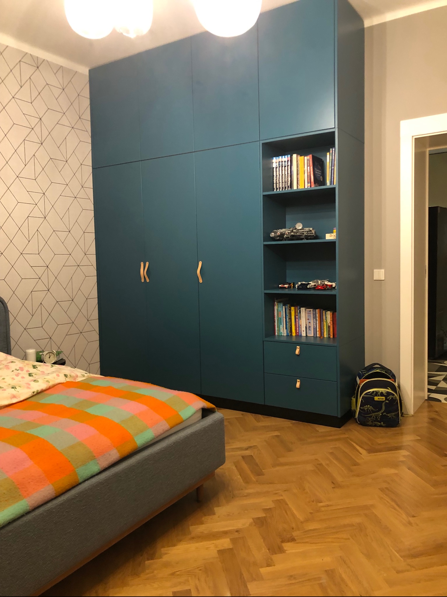 Niebieska szafa wnękowa z półkami na książki i zabawkami w pokoju dziecięcym, widoczne skórzane uchwyty, parkiet w jodełkę i plecak szkolny obok.