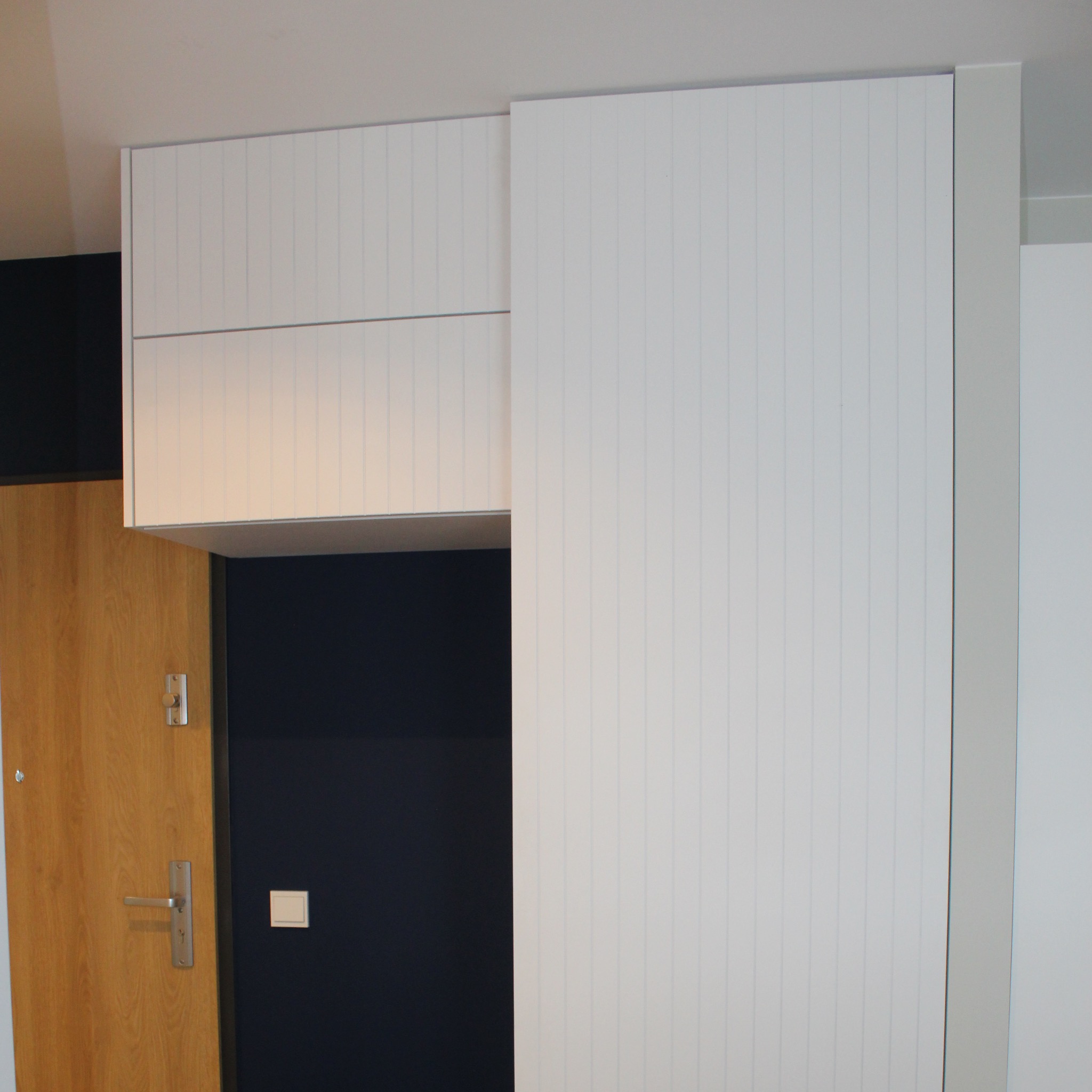 Biała szafa z pionowymi żłobieniami i górnymi szafkami zamontowana w przedpokoju obok drewnianych drzwi wejściowych i granatowej ściany.