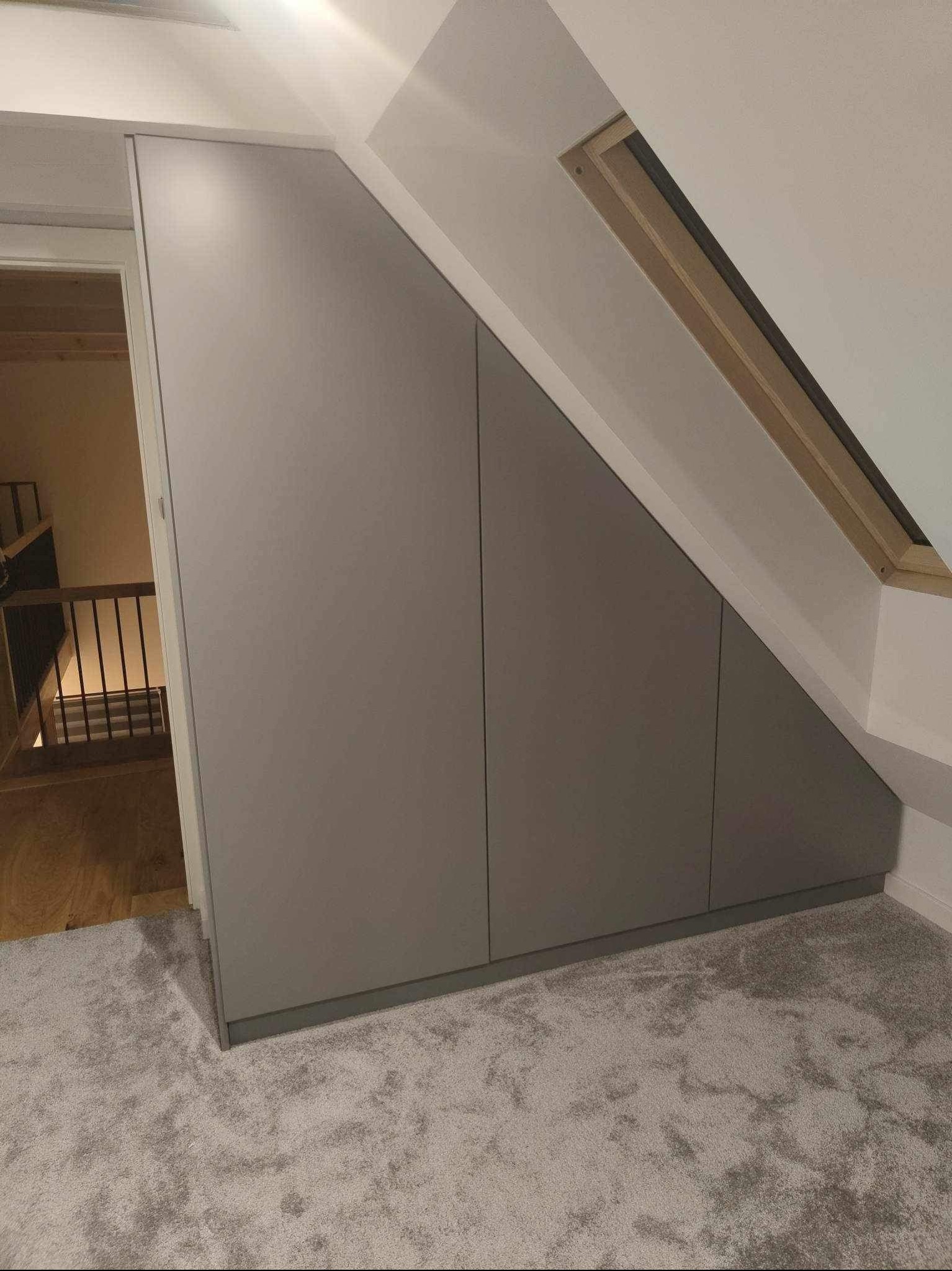 Szara zabudowa pod skosem na poddaszu z widocznym oknem dachowym i fragmentem schodów z drewnianą balustradą.