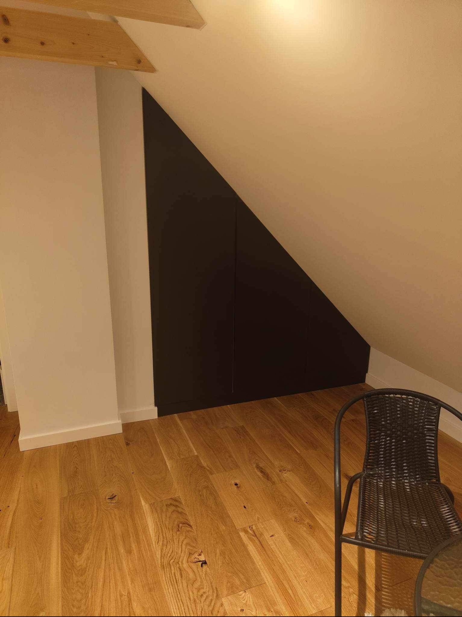 Ciemna, dwudrzwiowa szafa wnękowa pod skosem dachu, drewniana podłoga, krzesło z rattanu w rogu pokoju.