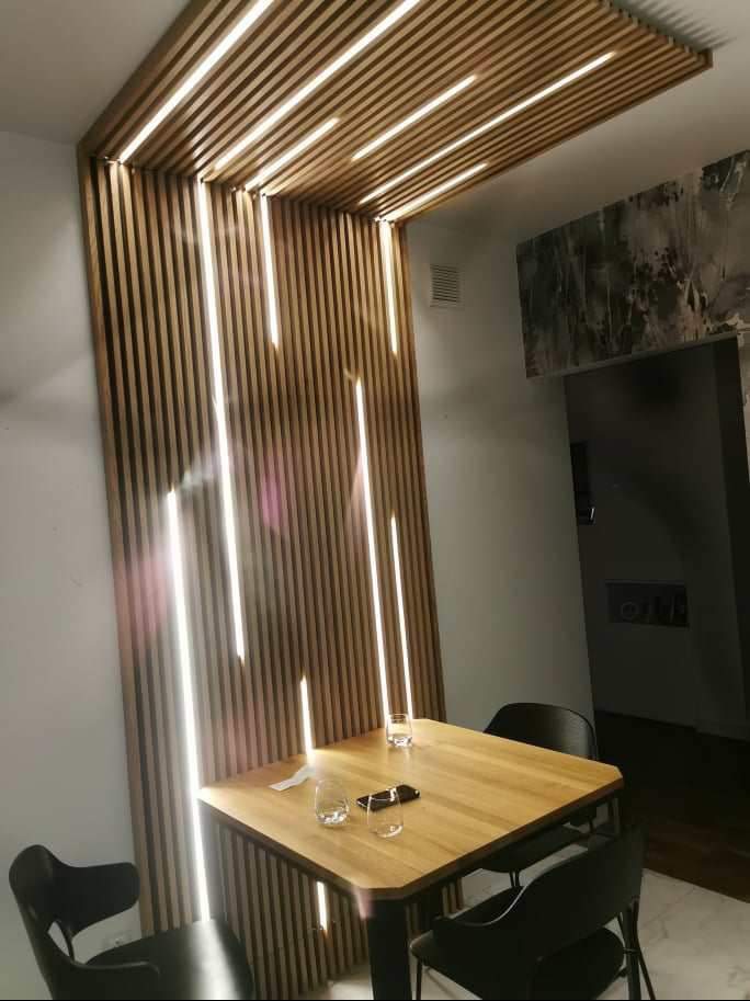 Drewniana ściana działowa z wbudowanym oświetleniem LED, tworząca dekoracyjny element w jadalni z kwadratowym stołem i czarnymi krzesłami.