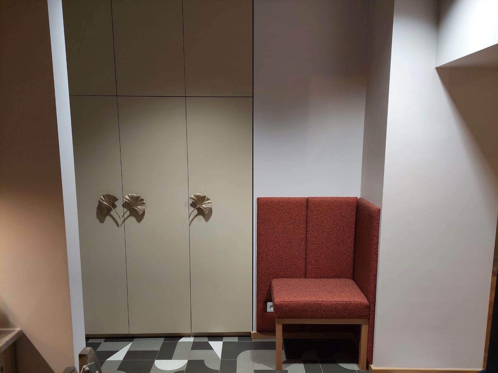 Beżowa szafa z trzema drzwiami, ozdobnymi uchwytami w kształcie liści ginkgo, stoi obok tapicerowanego siedziska w kolorze ceglastym z wysokim oparciem, na podłodze wzór geometryczny.