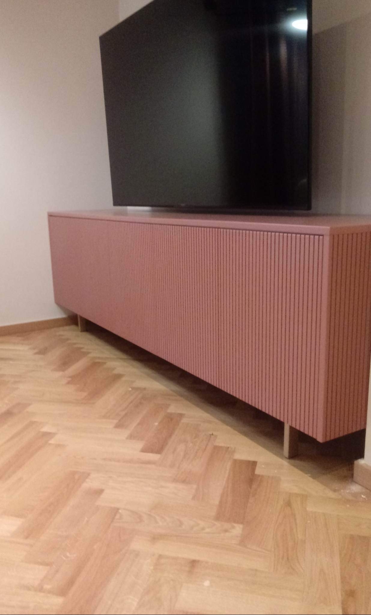 Różowa szafka RTV na metalowych nóżkach z ryflowanym frontem pod telewizorem na parkiecie ułożonym w jodełkę.