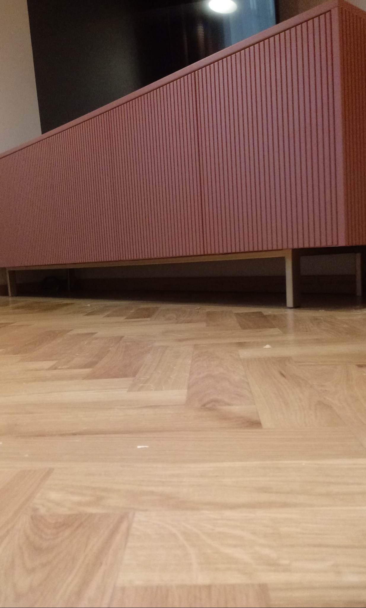 Długa, niska komoda w kolorze terakoty z ryflowanymi frontami, osadzona na metalowych nóżkach, na tle drewnianej podłogi parkietowej.