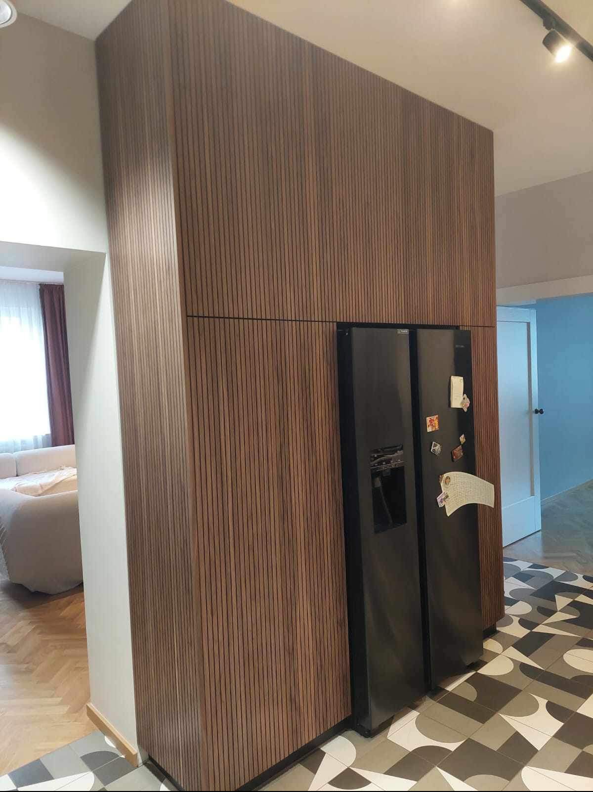 Panel ścienny z pionowych listew drewnianych w kolorze orzecha, zabudowujący lodówkę w nowoczesnym wnętrzu z wzorzystą podłogą.