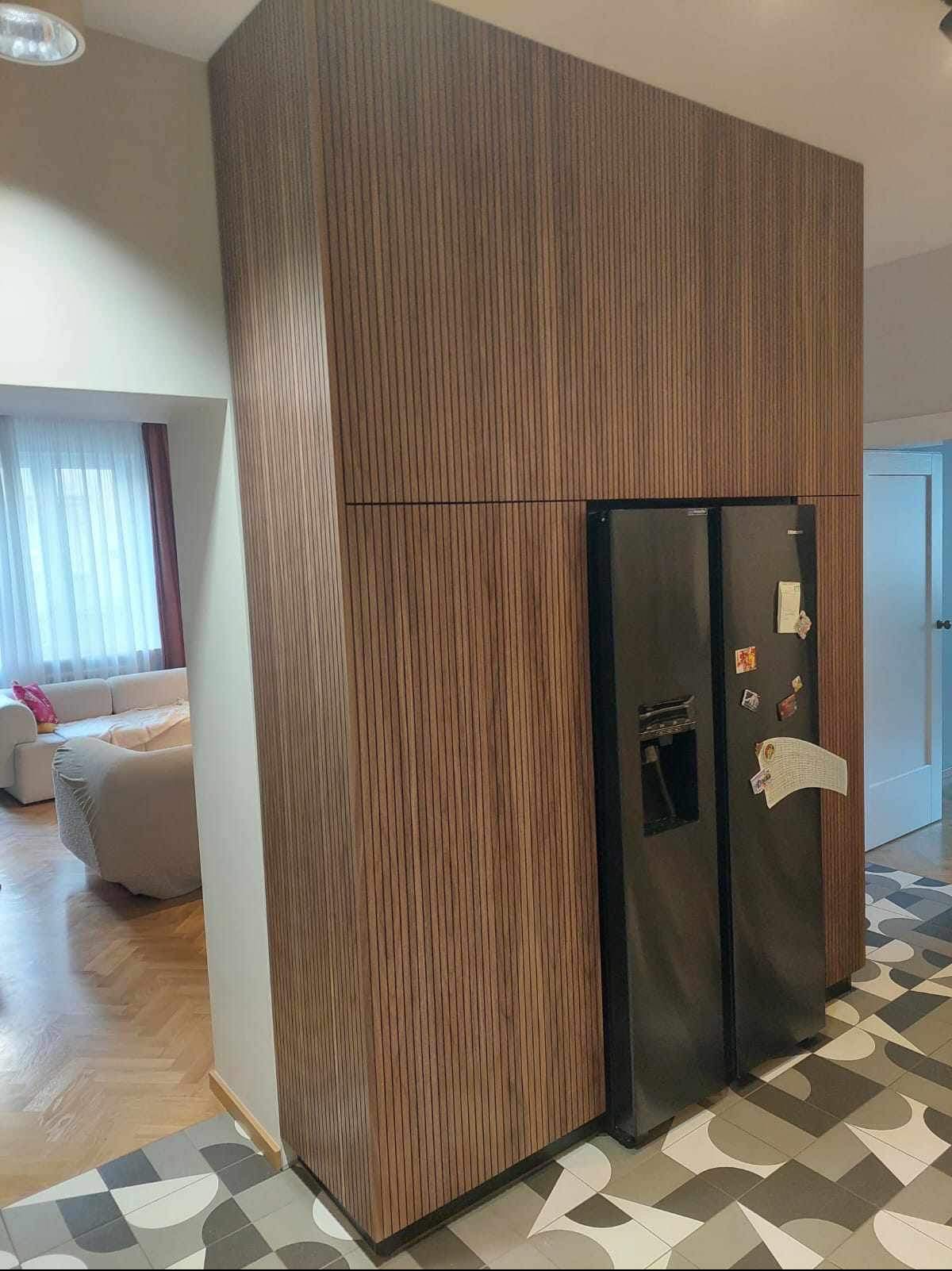 Nowoczesna kuchnia z zabudową z paneli drewnianych, w której wkomponowano lodówkę side-by-side; w tle widoczny salon z jasną sofą.