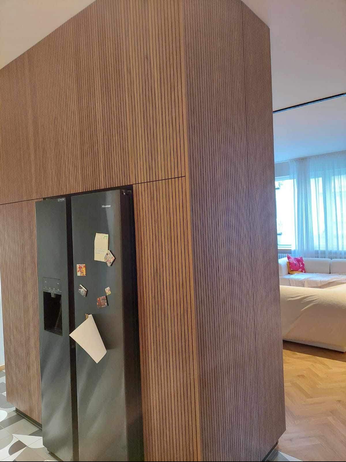 Nowoczesna kuchnia z zabudową z paneli drewnianych, wkomponowana czarna lodówka, w tle widoczny salon z sofą.