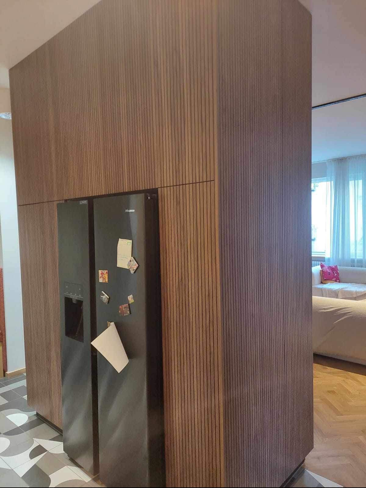 Nowoczesna zabudowa kuchenna z pionowych listew drewnianych, integrująca lodówkę z magnesami i notatkami, widoczna z perspektywy salonu.