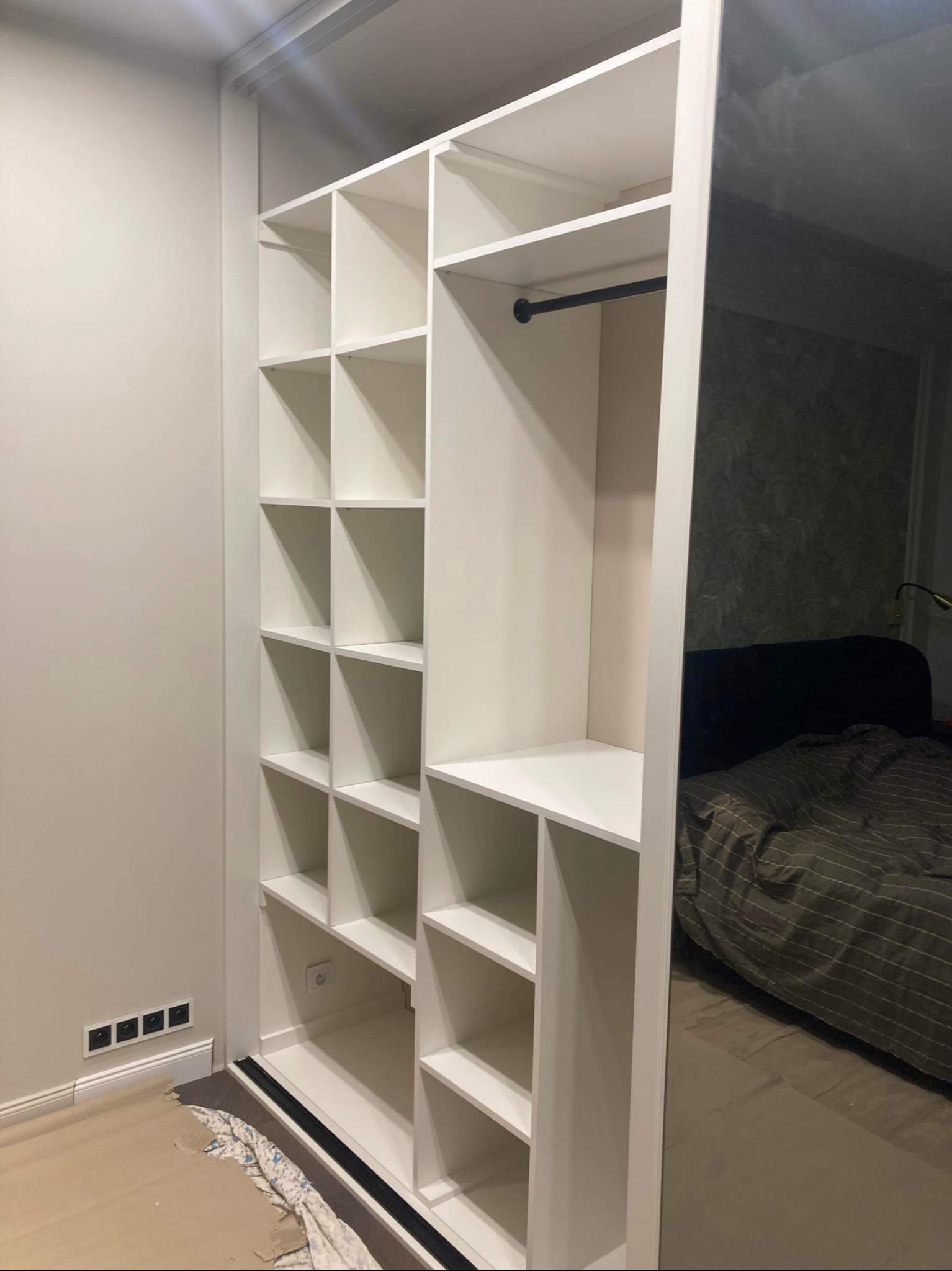 Biała szafa wnękowa z półkami i drążkiem na ubrania, widoczne gniazdka elektryczne na ścianie obok i odbicie sypialni w lustrzanych drzwiach.