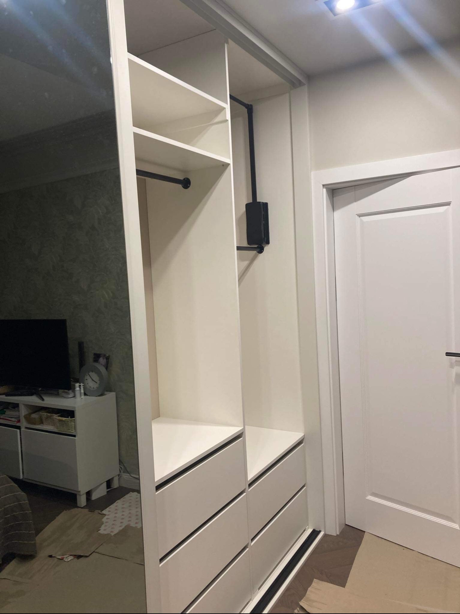 Biała szafa z szufladami i półkami, zamontowana w pokoju, widoczna instalacja elektryczna, lustrzane drzwi przesuwne po lewej stronie.