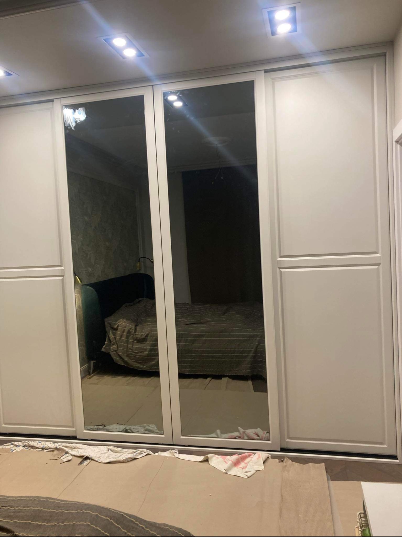 Biała szafa przesuwna z lustrami w trakcie montażu, zabezpieczona kartonem na podłodze, widoczne odblaski sufitowych lamp i fragment sypialni w lustrach.