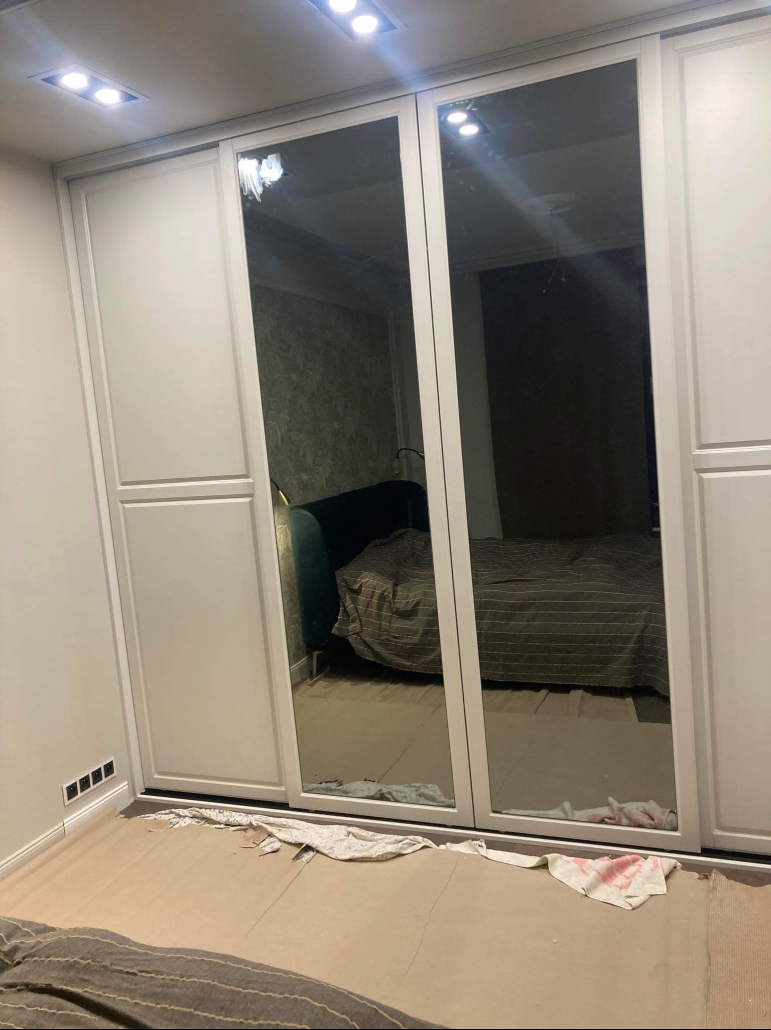Biała szafa przesuwna z lustrami wbudowanymi w drzwi, odbijająca fragment sypialni z łóżkiem i zielonym zagłówkiem. Na podłodze widoczne zabezpieczenia i fragmenty materiału.