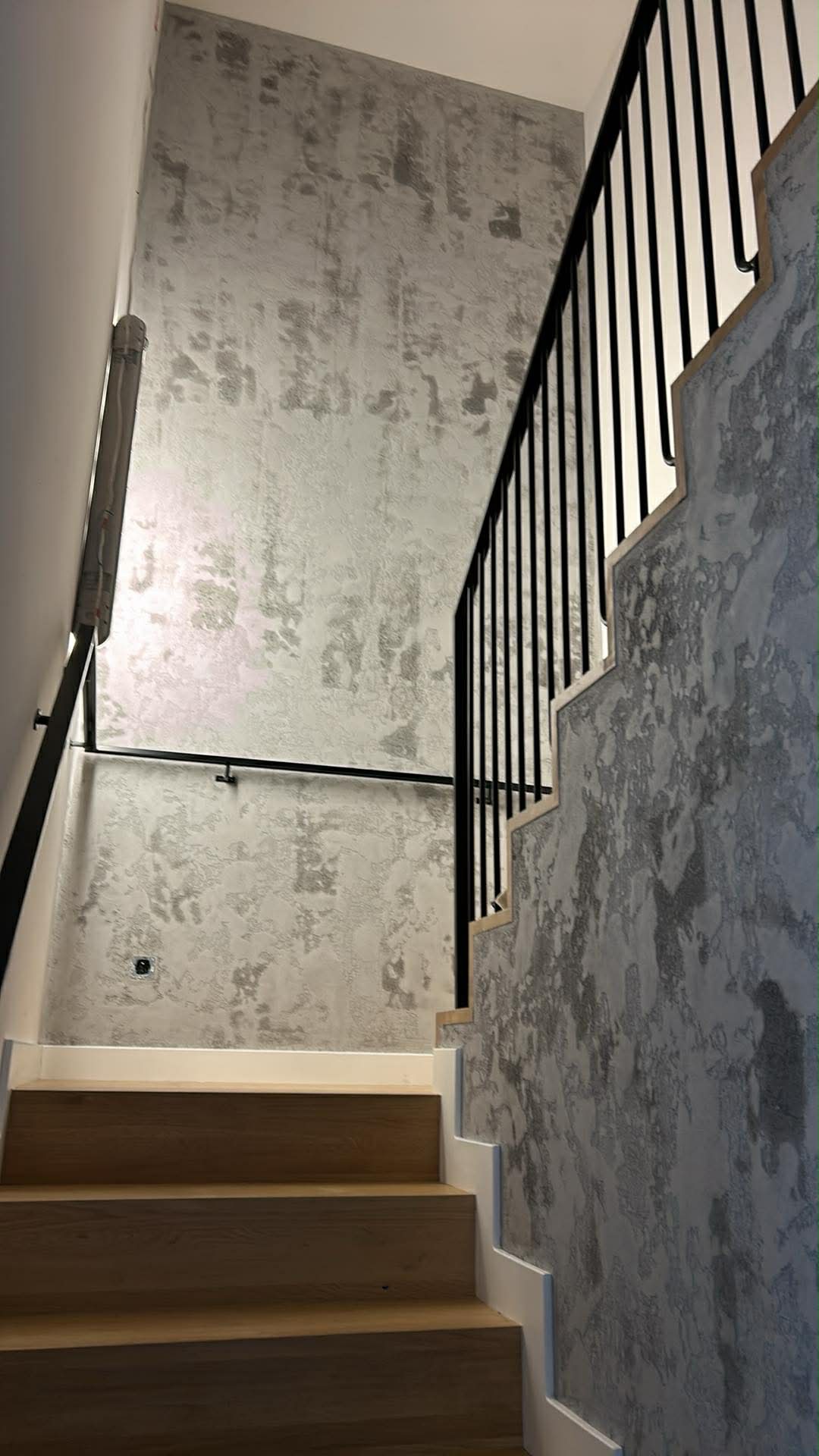 Schody z drewnianymi stopniami i ścianami wykończonymi dekoracyjną, szarą strukturą. Czarna, metalowa balustrada. Nowoczesny design klatki schodowej.