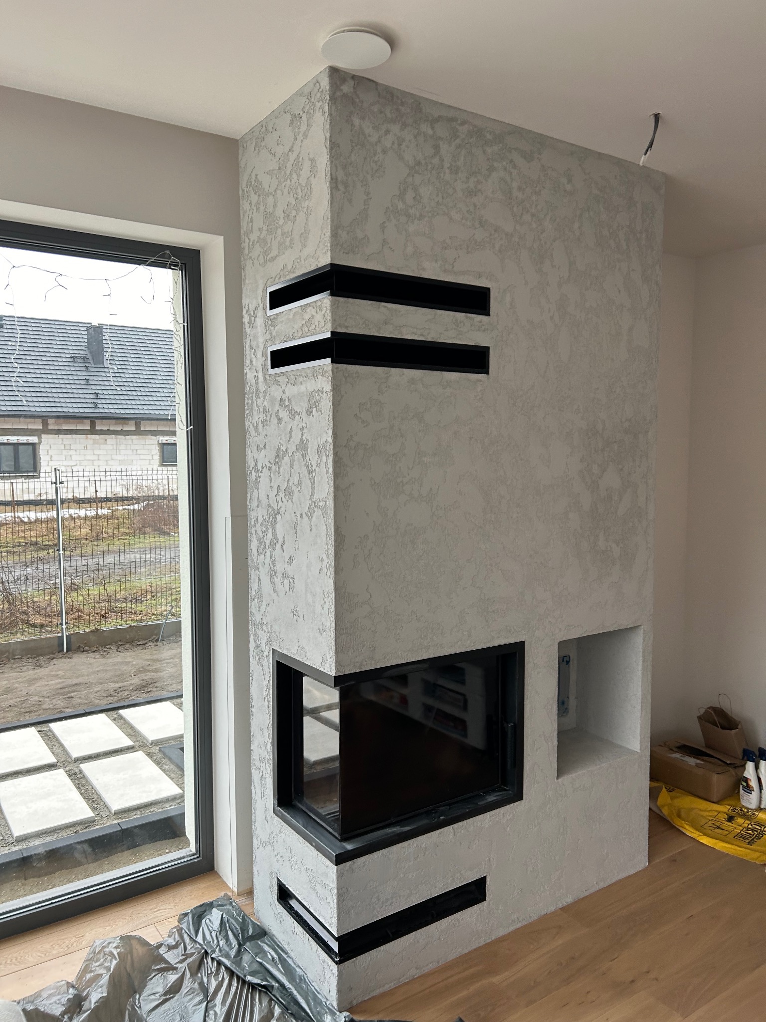 Wykończony kominek z szarą, dekoracyjną strukturą, czarnymi akcentami i wkładem kominkowym. Widoczne fragmenty podłogi i okna.