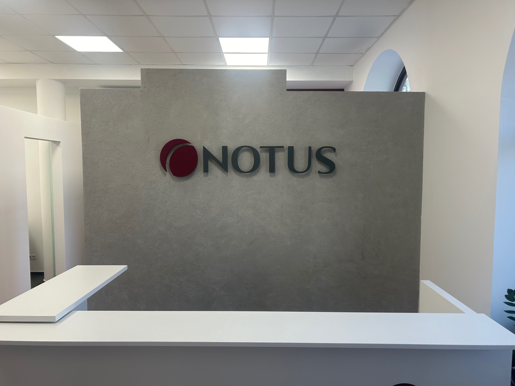 Recepcja biura z szarą ścianą o fakturze betonu, na której umieszczono logo firmy 'NOTUS' w kolorze czerwono-szarym. Białe elementy recepcji i oświetlenie sufitowe.