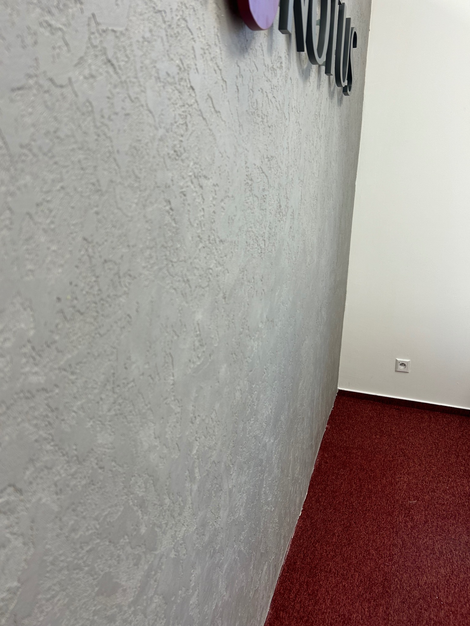 Szara ściana z dekoracyjną strukturą, widok z dołu, z fragmentem napisu i gniazdkiem elektrycznym. Czerwona wykładzina podłogowa.