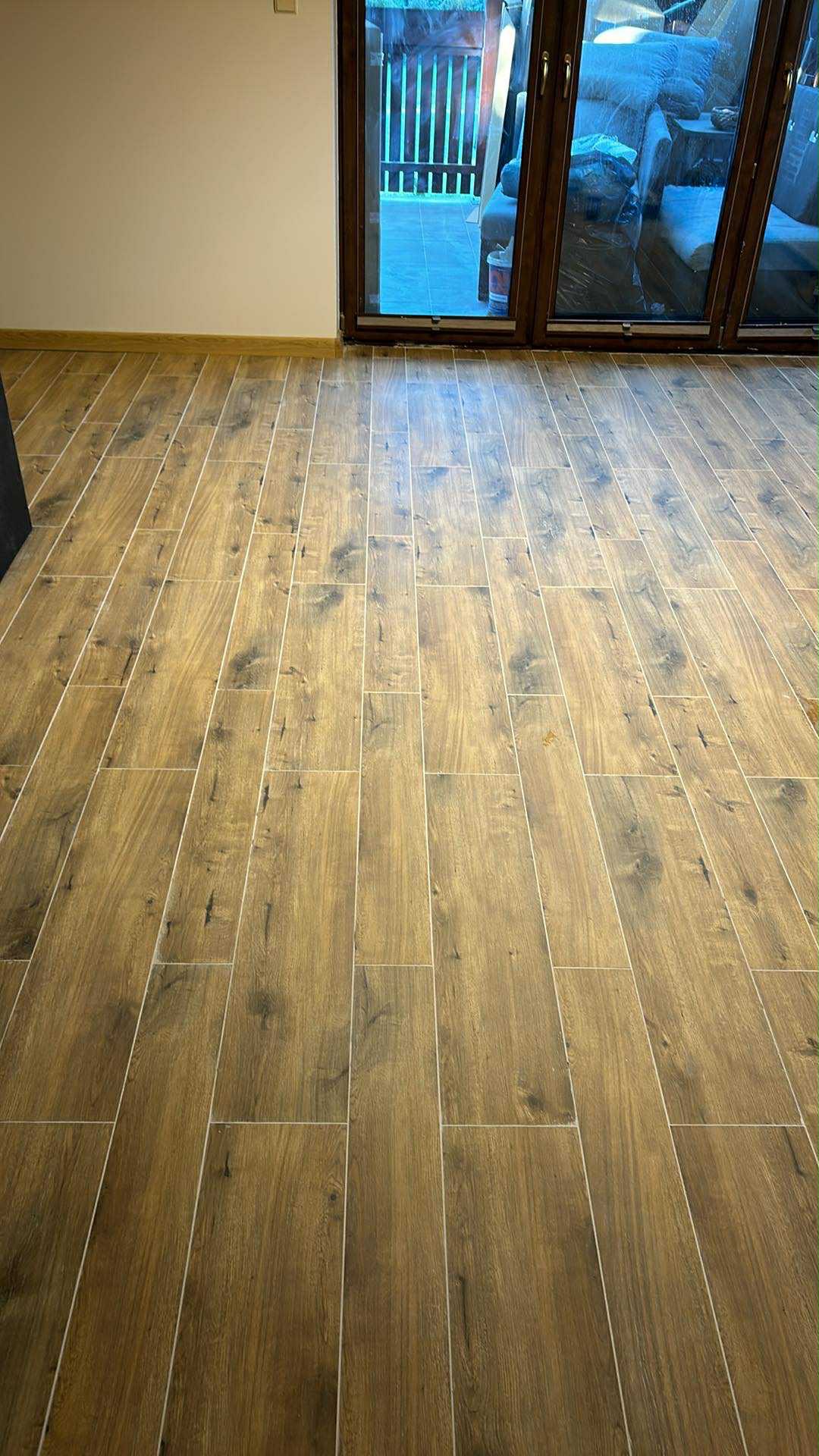 Ułożona podłoga z płytek imitujących drewno, widok z góry, jasne fugi, duże okno balkonowe w tle, ściana w neutralnym kolorze. Wykończenie podłogi.