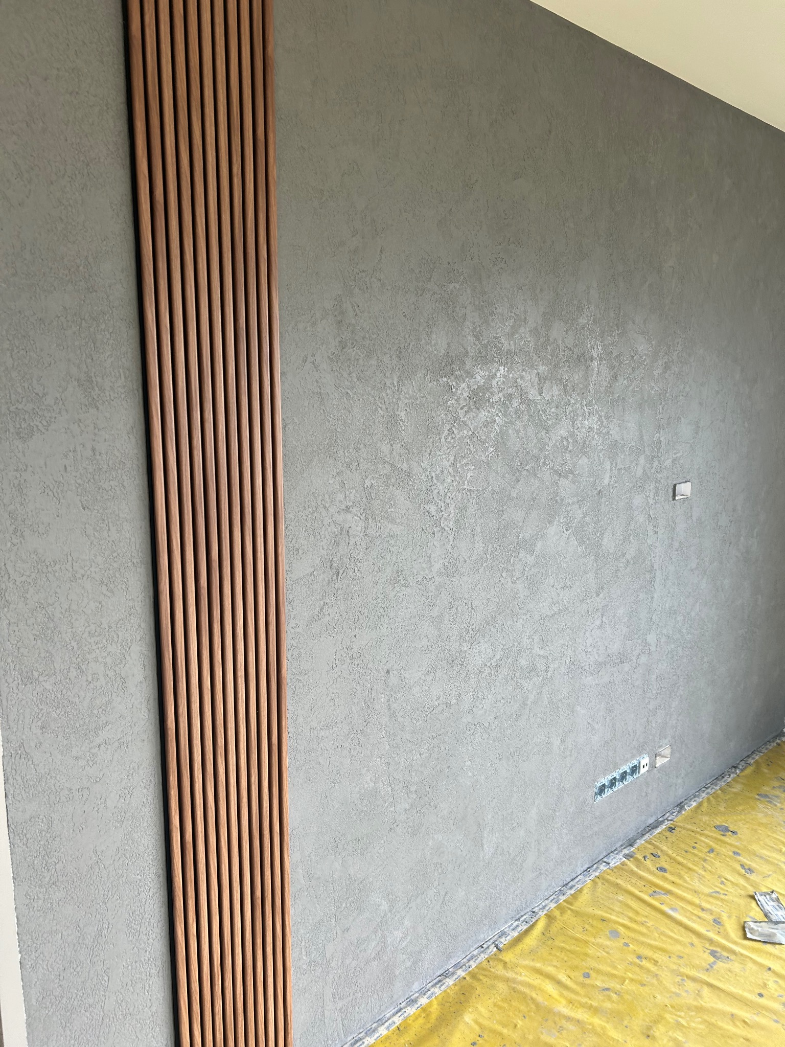Szara ściana z dekoracyjną, pionową listwą z drewna. Widoczne gniazdka elektryczne i zabezpieczona folią podłoga. Wykończenie wnętrza w nowoczesnym stylu.