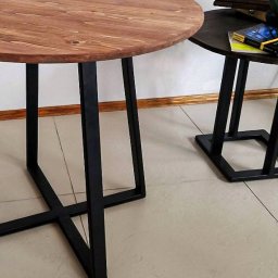 LoftOn - Okrągły stolik kawowy z blatem z naturalnego drewna i czarnymi, metalowymi nogami o geometrycznym kształcie. W tle drugi, mniejszy stolik z ciemnym blatem i książkami.