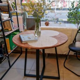 LoftOn - Okrągły stolik z blatem z jasnego drewna i czarnymi metalowymi nogami, na blacie wazon z żółtymi kwiatami i książka, w tle witryna sklepu.