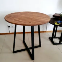 Okrągły stół z blatem z jasnego drewna i czarnymi, metalowymi nogami w kształcie litery X, widoczny na jasnej, kafelkowanej podłodze, z gniazdkiem elektrycznym w tle.