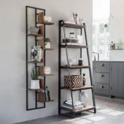 Dwie metalowe półki z drewnianymi blatami w stylu industrialnym, jedna przymocowana do ściany, druga w formie drabiny, eksponujące dekoracje, naczynia i książki w nowoczesnym wnętrzu.