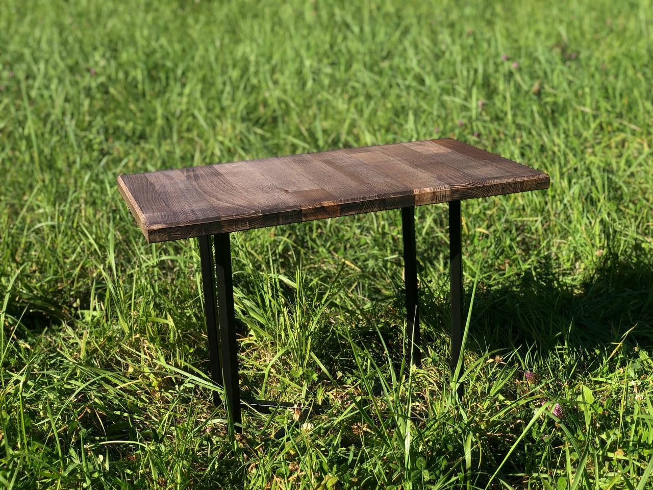 Niewielki, prostokątny stolik z blatem z ciemnego drewna i czarnymi, metalowymi nogami, ustawiony na trawie w plenerze.
