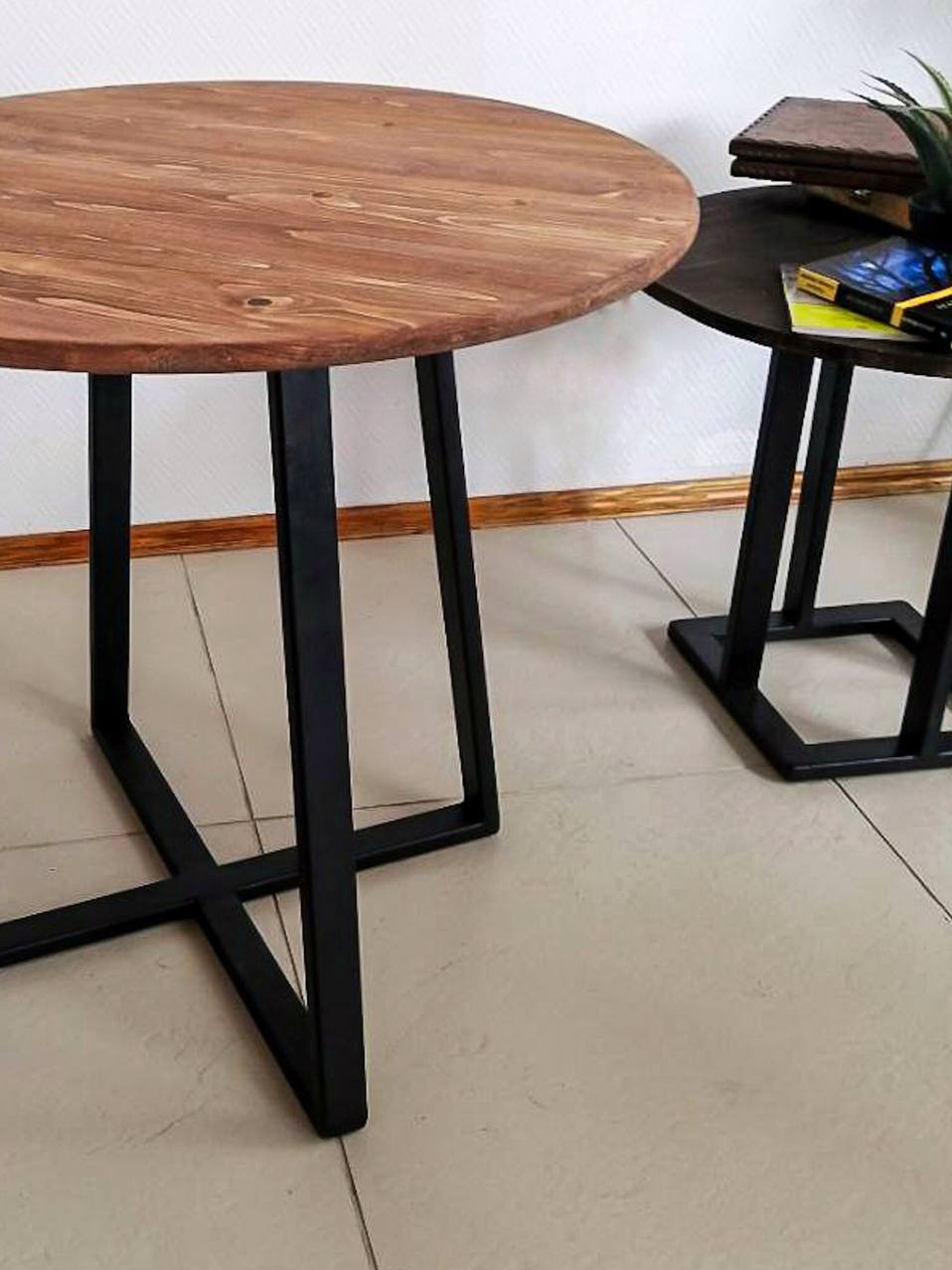 Okrągły stolik kawowy z blatem z naturalnego drewna i czarnymi, metalowymi nogami o geometrycznym kształcie. W tle drugi, mniejszy stolik z ciemnym blatem i książkami.