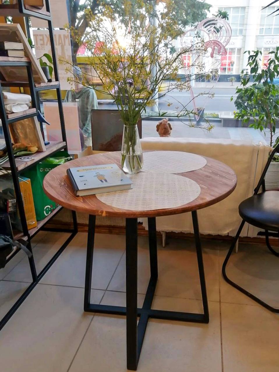 Okrągły stolik z blatem z jasnego drewna i czarnymi metalowymi nogami, na blacie wazon z żółtymi kwiatami i książka, w tle witryna sklepu.