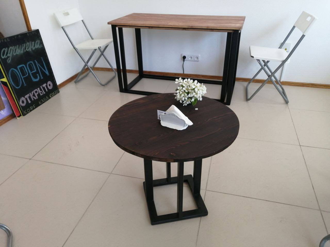 Okrągły, ciemny stolik kawowy z drewnianym blatem i czarną, metalową podstawą w kawiarni, obok tablica z napisem 'OPEN' i serwetki na blacie.