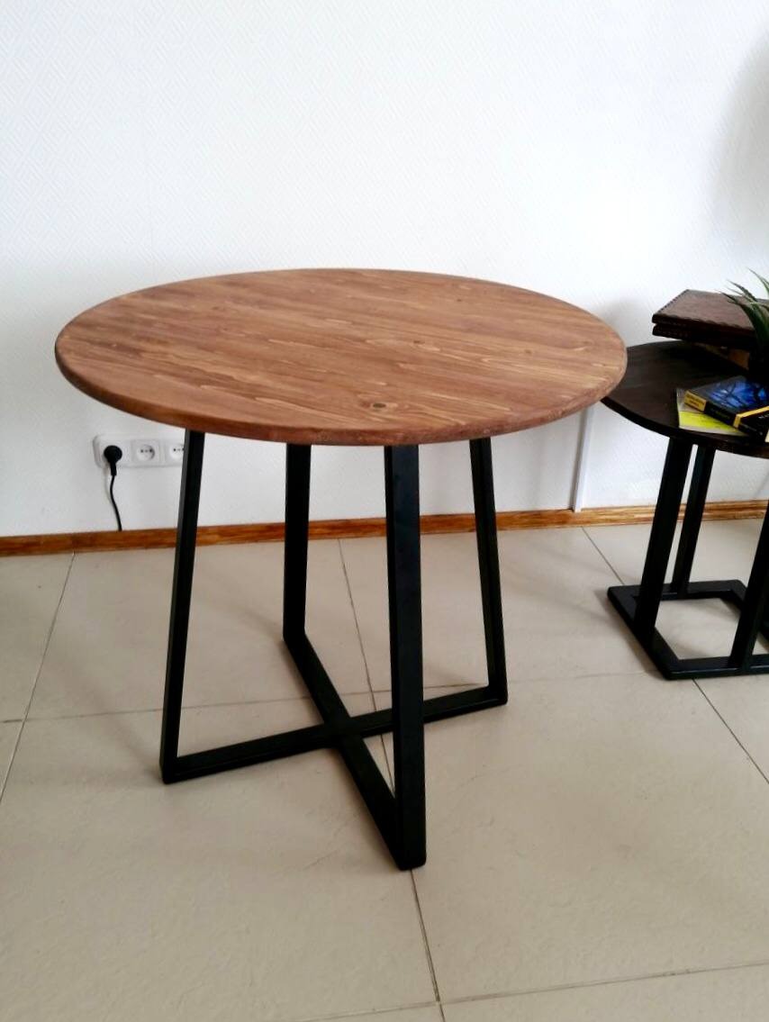 Okrągły stół z blatem z jasnego drewna i czarnymi, metalowymi nogami w kształcie litery X, widoczny na jasnej, kafelkowanej podłodze, z gniazdkiem elektrycznym w tle.