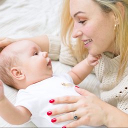 Alicja Drożyńska - Matka z blond włosami i czerwonym manicure patrzy z uśmiechem na leżącego niemowlaka w białym body z aplikacją, na kremowym tle.