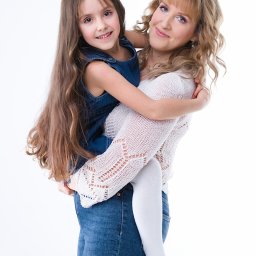 Alicja Drożyńska - Uśmiechnięta mama w białym swetrze tuli córkę w dżinsowej sukience na białym tle. Profesjonalne oświetlenie podkreśla naturalne piękno modelek.