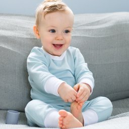 Alicja Drożyńska - Uśmiechnięty niemowlak w błękitnym ubranku siedzi na szarym siedzisku, trzymając stópkę w dłoniach, z widocznym fragmentem szarego kubeczka obok.