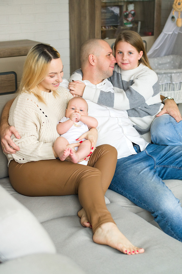 Szczęśliwa rodzina siedzi na szarej sofie: mama trzyma noworodka, tata obejmuje starszą córkę, w tle widoczny regał i dziecięce łóżeczko.