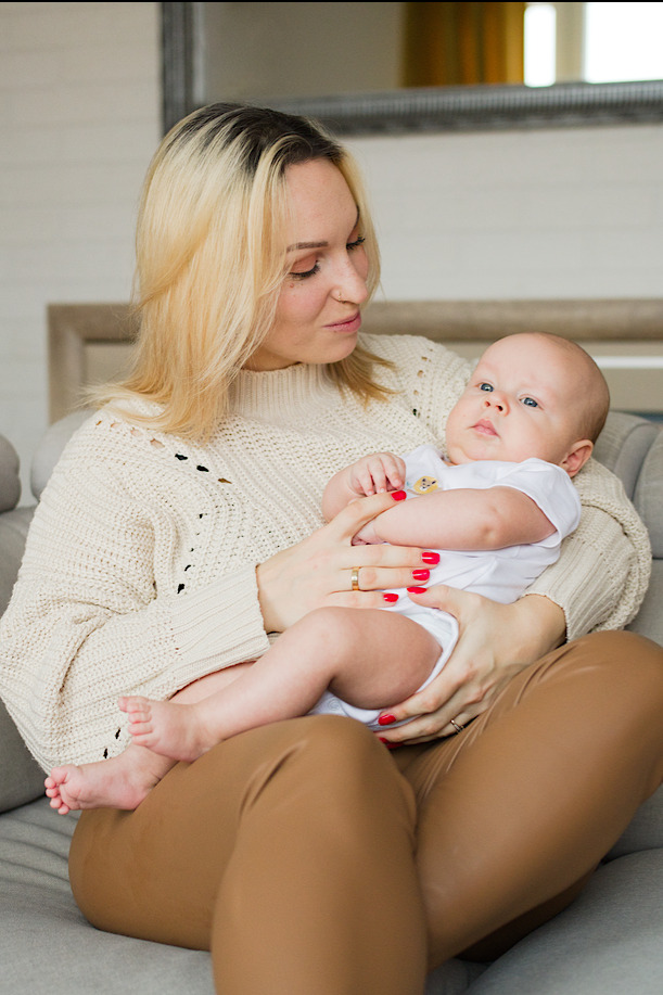 Mama z blond pasemkami, w kremowym swetrze, trzyma na kolanach niemowlę w białym body na szarym tle kanapy, z widocznymi małymi stopkami dziecka i czerwonym manicure matki.