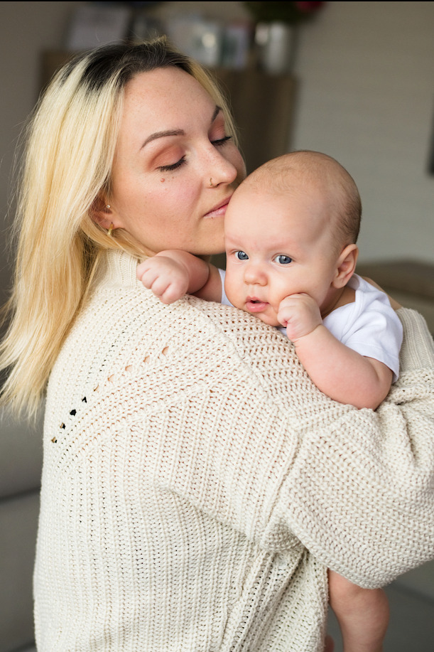 Blondwłosa mama w kremowym swetrze całuje w głowę niemowlę o jasnej cerze i niebieskich oczach, które opiera policzek na jej ramieniu.