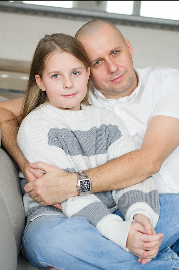 Portret ojca obejmującego córkę w swetrze w paski, oboje siedzą na kanapie w jasnym wnętrzu.